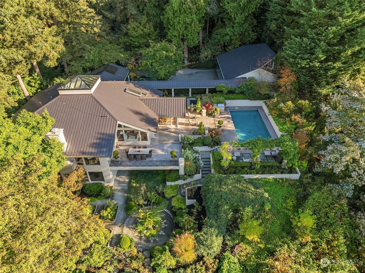 An aerial view showcases a modern house with a swimming pool and extensive landscaping, surrounded by a dense forest.