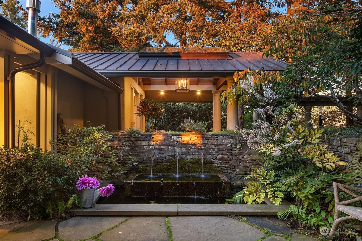 A tranquil outdoor courtyard features a stone fountain, lush landscaping, and a partially visible house with warm lighting.