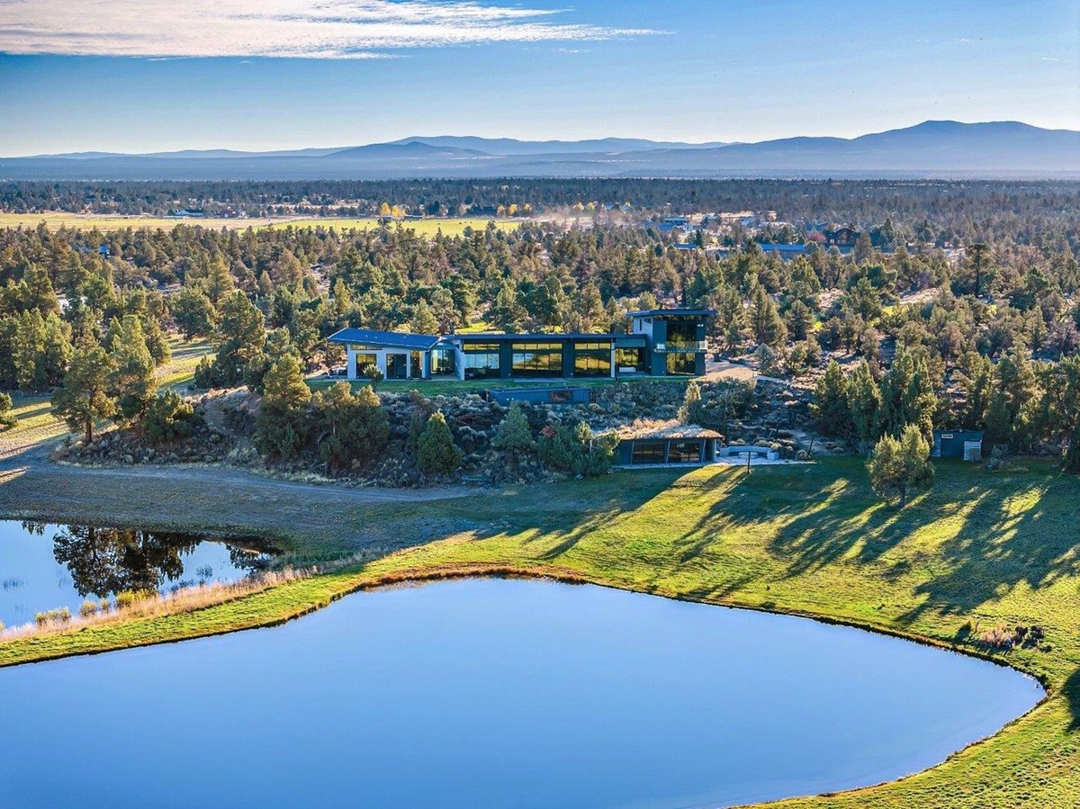 Modern house surrounded by trees, located near a reflective pond with distant mountains visible under a clear sky.