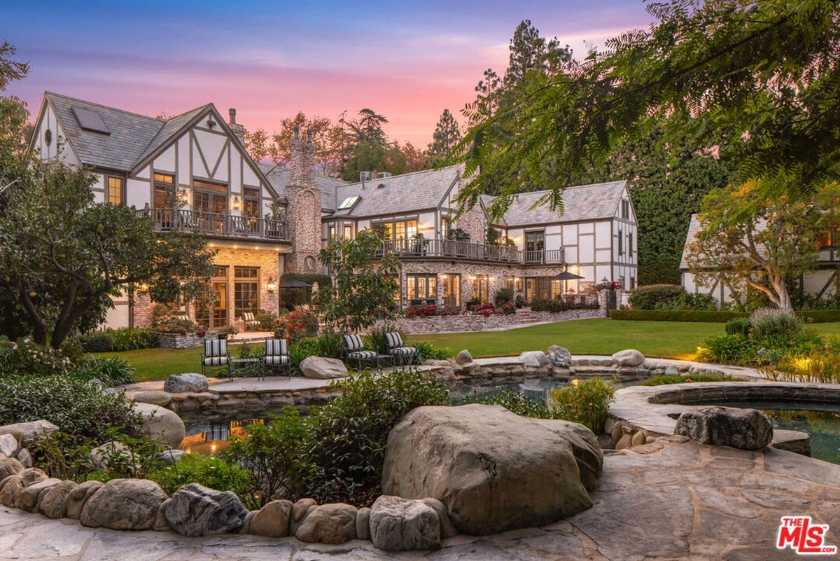 A majestic Tudor-style mansion, captured from a slightly elevated rear angle, showcases its expansive grounds and meticulously landscaped pool area. The image highlights the home's impressive size and the serene beauty of its outdoor space, creating a picture of luxurious living.