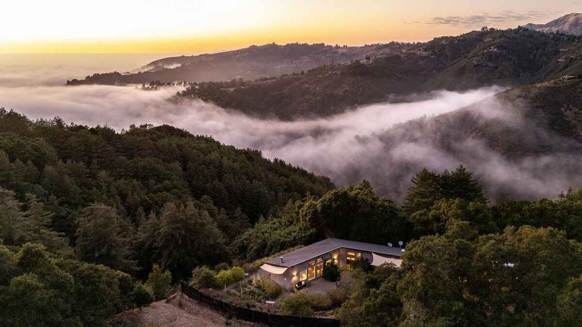 Aerial view of a house nestled in a lush, fog-draped mountain valley at sunset.