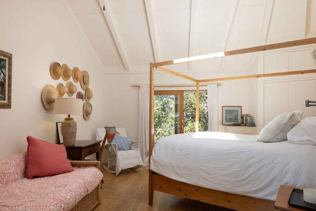 Wooden four-poster bed dominates a sunlit bedroom with a vaulted ceiling, showcasing a collection of straw hats on the wall.