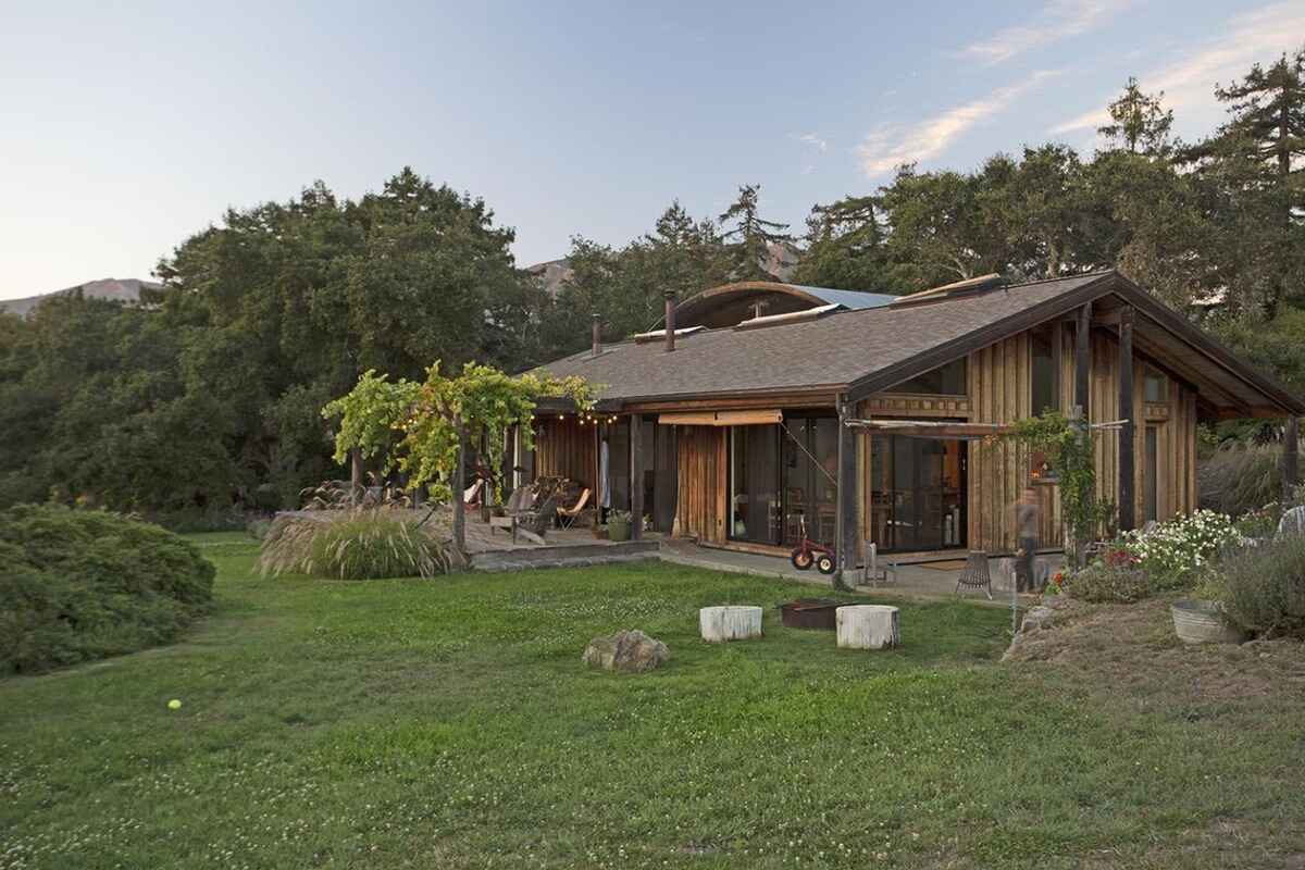 Long, low-slung house with wood siding and a large lawn sits against a backdrop of trees and hills.