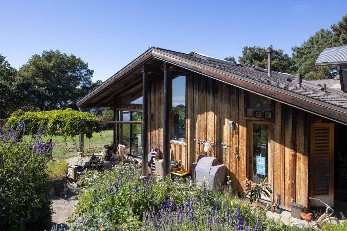 Rustic wooden house with a steeply pitched roof and large windows is surrounded by lush landscaping.