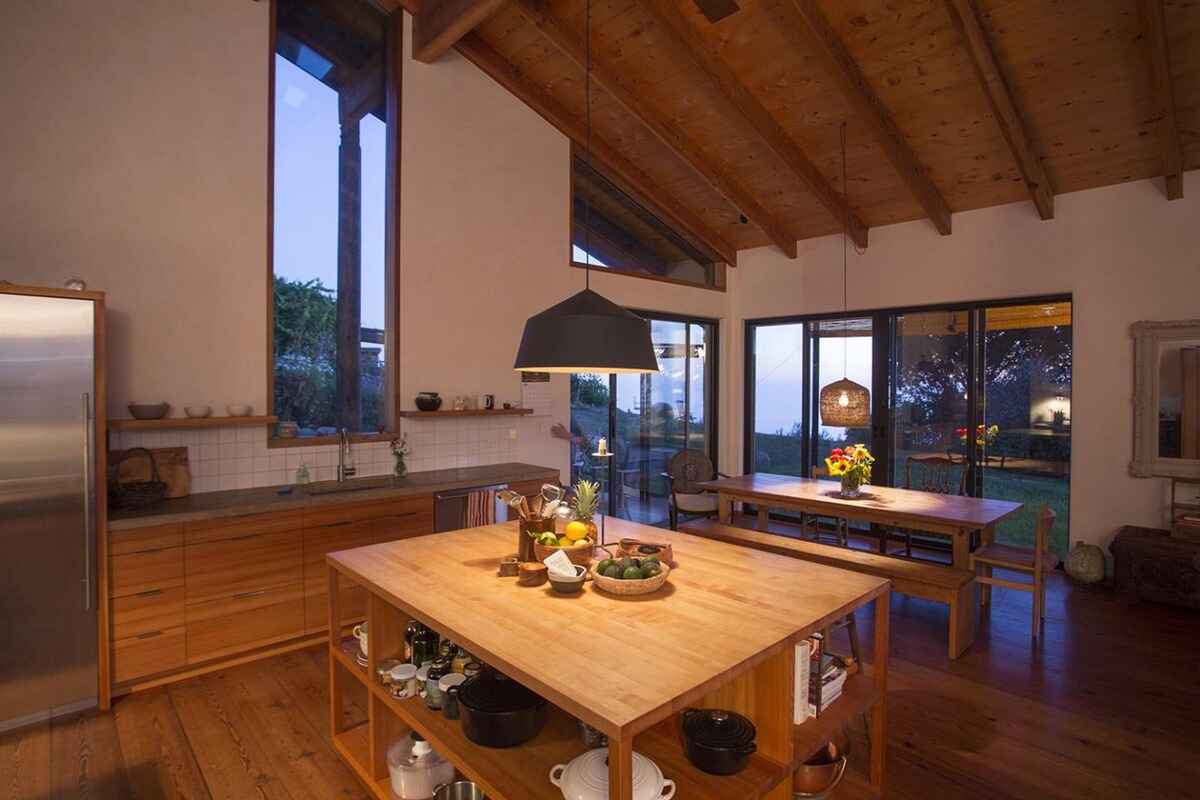 Modern kitchen with wood cabinets, a large island, and a dining area, all illuminated by pendant lights and enjoying natural light from large windows.