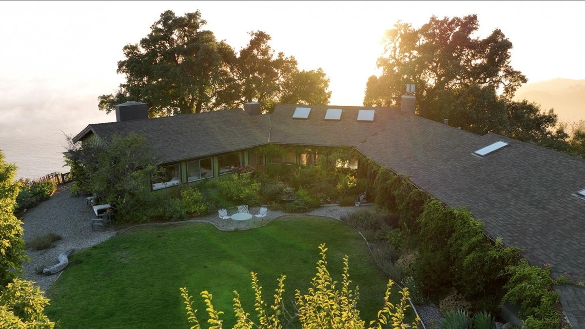 Sunlight illuminates a sprawling ranch-style home surrounded by a meticulously landscaped yard and garden, overlooking a distant ocean.