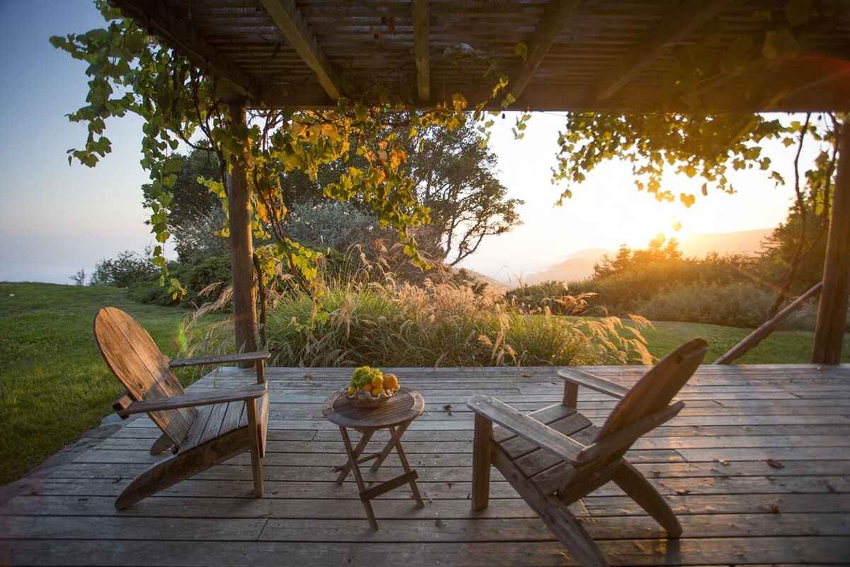 Two wooden Adirondack chairs flank a small table holding fruit on a deck overlooking a sunset view, all shaded by a pergola covered in vines.