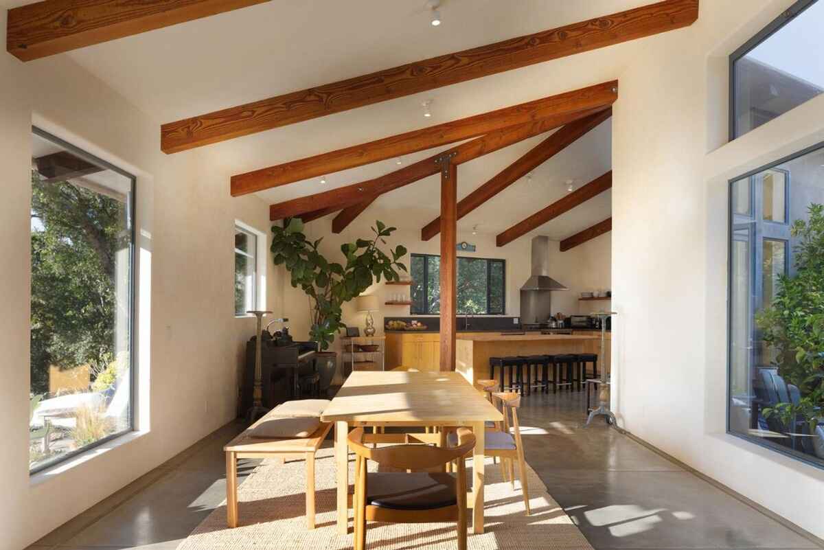 Dining area with a long wooden table and benches, situated in a sunlit room with exposed wooden beams and large windows.