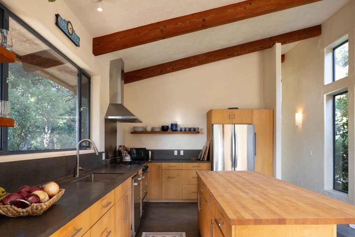 Modern kitchen with light wood cabinets, a large island with a butcher block top, and a stainless steel range hood, all under a wood-beamed ceiling.