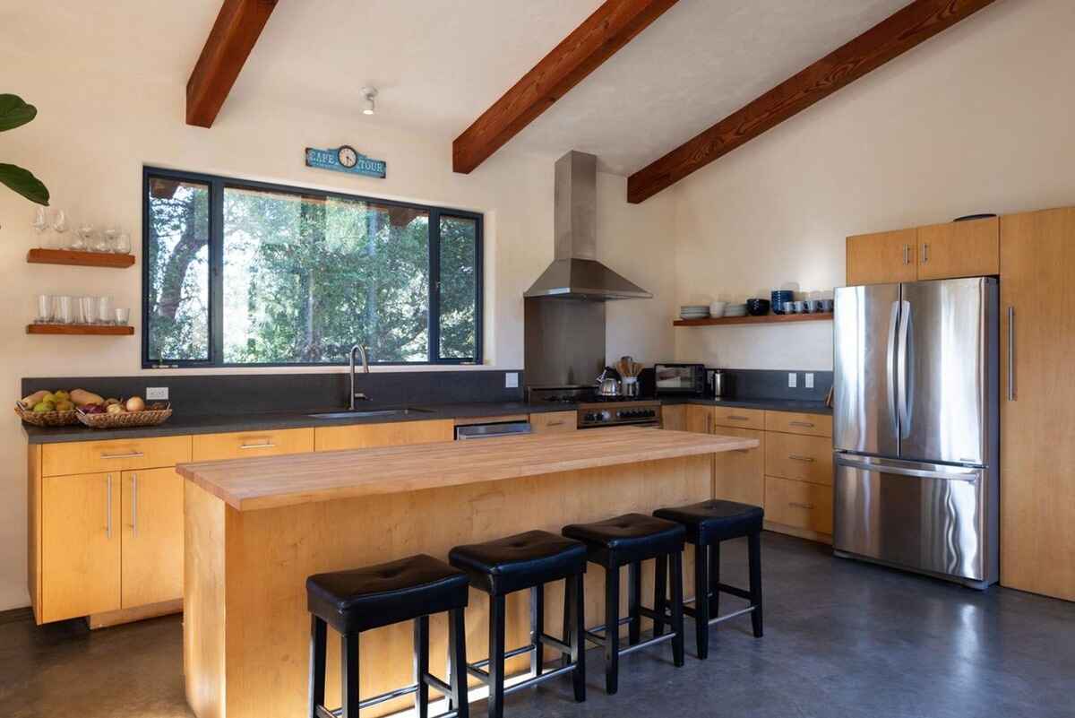 Modern kitchen with light wood cabinets, a large island with a butcher block top, and a stainless steel refrigerator, all under a wood-beamed ceiling.