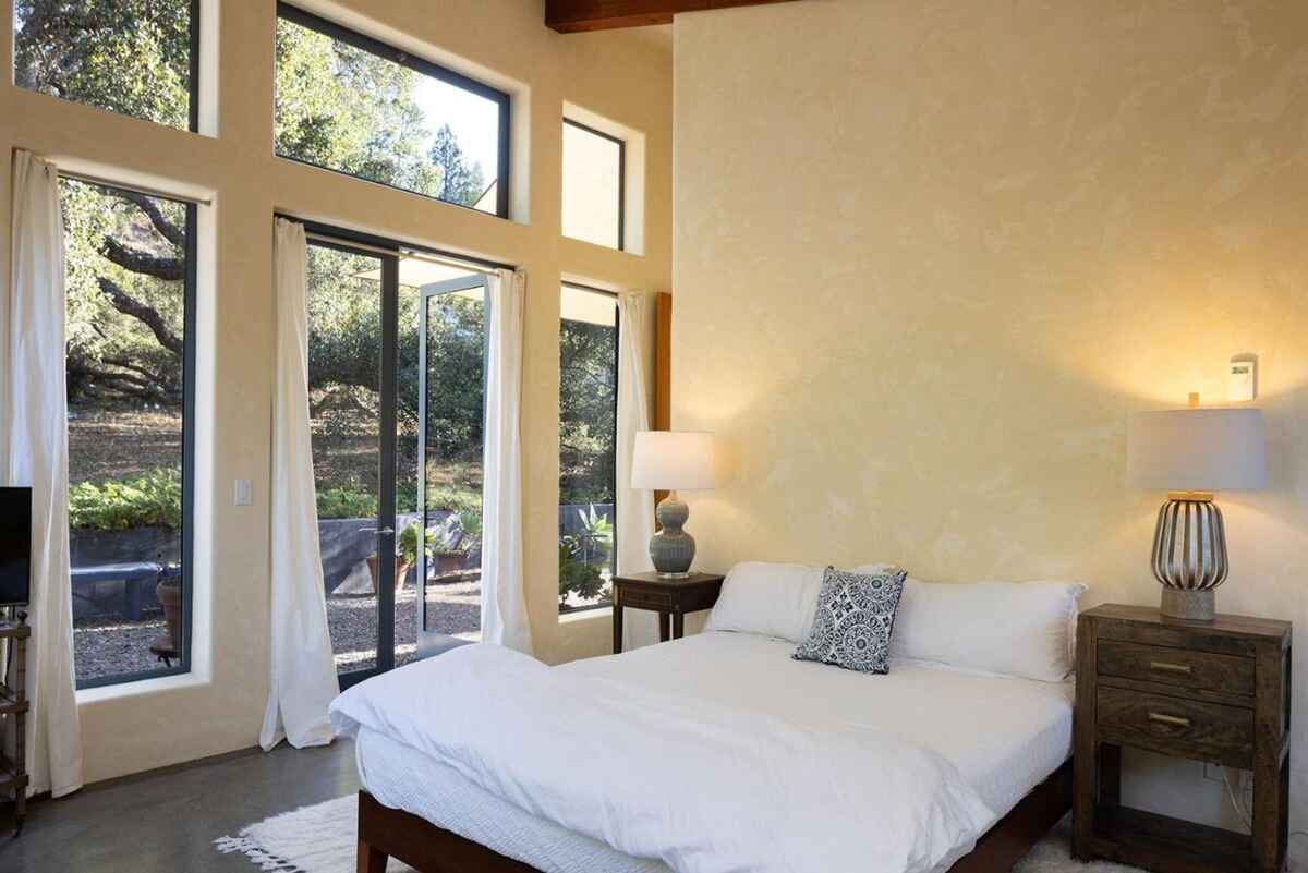 Bedroom with a bed dressed in white linens, situated against a textured wall and featuring large windows offering views of trees and greenery.