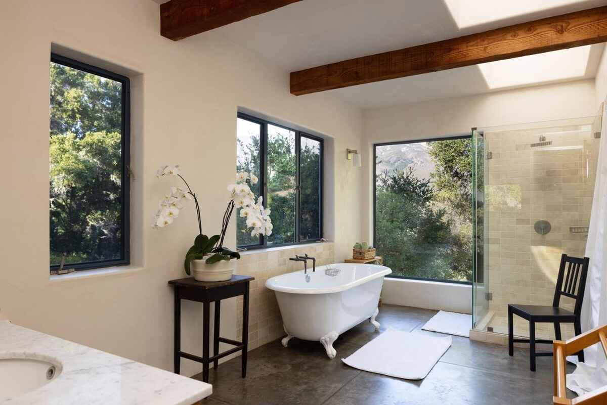 Bathroom with a clawfoot tub, a walk-in shower, and multiple windows offering views of nature, all under wood beams.