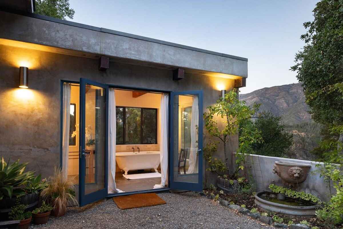 Outdoor view of a bathroom with a freestanding tub, accessed through French doors from a patio area, all set against a backdrop of hills and greenery.