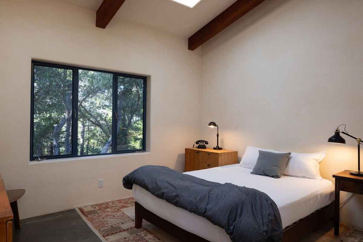 Bedroom with a bed featuring a dark gray duvet, situated near a window offering a view of trees, all under a wood-beamed ceiling.