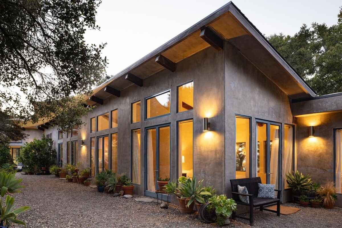 Long, low-slung house with stucco exterior and large windows, illuminated at dusk, sits amidst landscaping and trees.