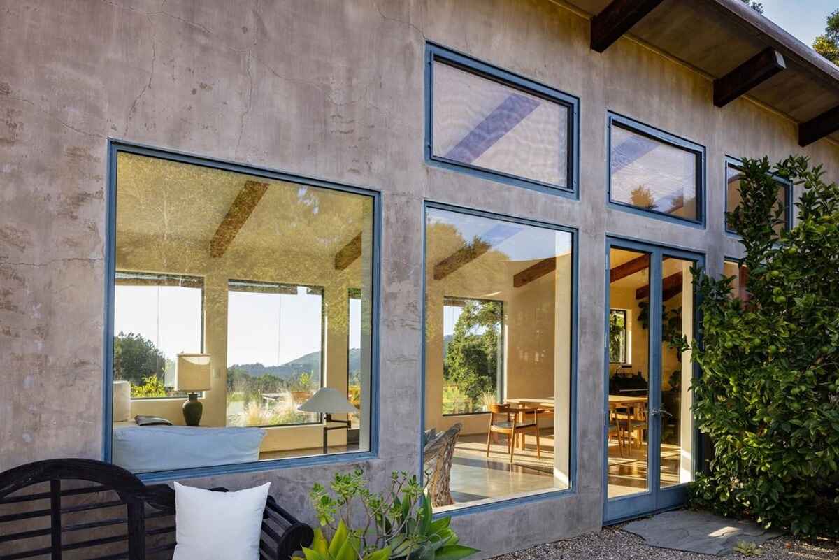 Large windows showcasing an interior living space and outdoor views are featured on a stucco exterior wall of a home.