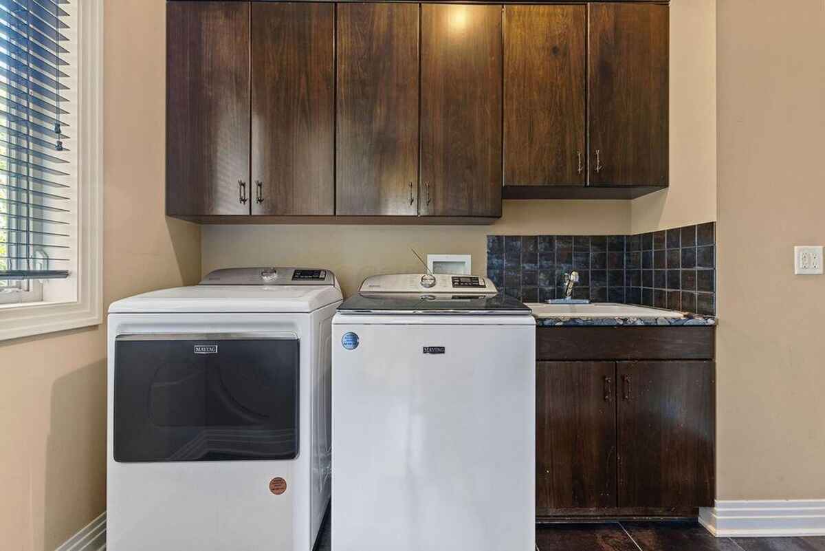 Laundry room showcases a washer and dryer set below dark wood cabinets, with a utility sink and tile backsplash.