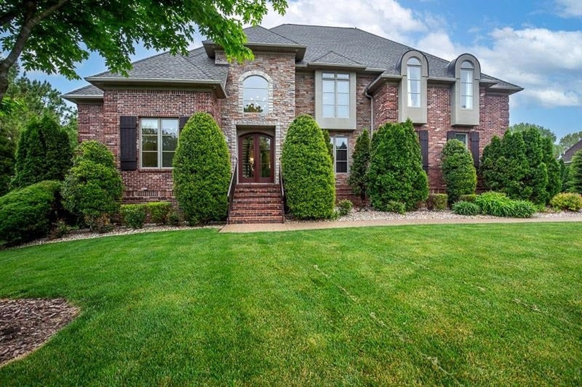  Brick house with manicured lawn, shrubs, and trees in front of the main entrance.