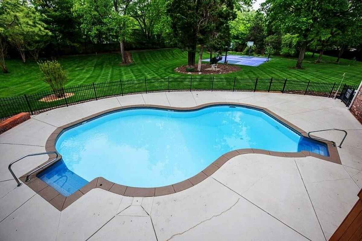 Curved swimming pool surrounded by concrete deck and fenced backyard with manicured landscaping.