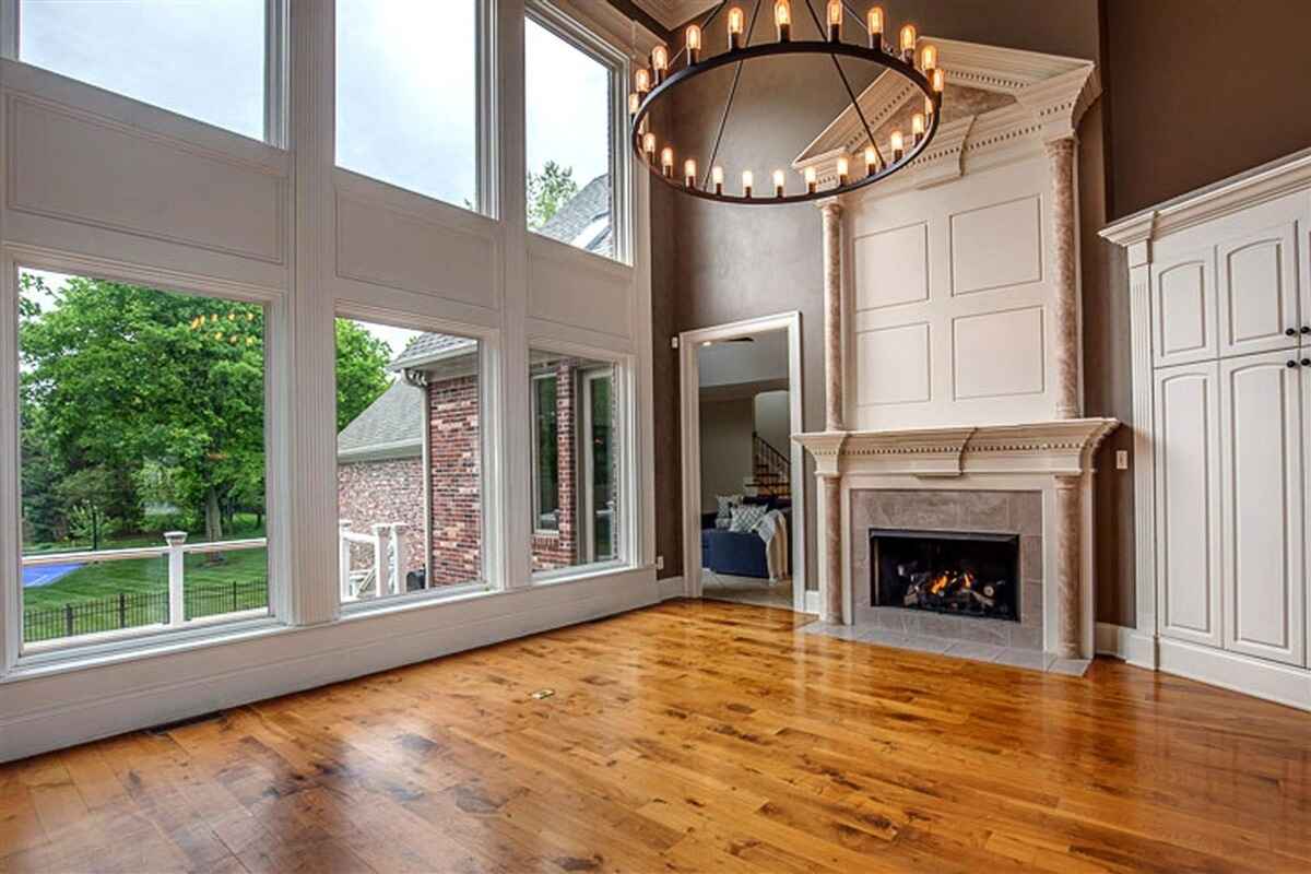 Living room corner showing large windows, fireplace, hardwood flooring, and a chandelier.