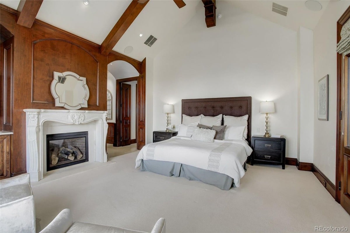 Architectural details such as exposed beams and rich wood paneling add to the grandeur of this master bedroom. The combination of a stately fireplace and comfortable bedding creates a space that is both elegant and relaxing.