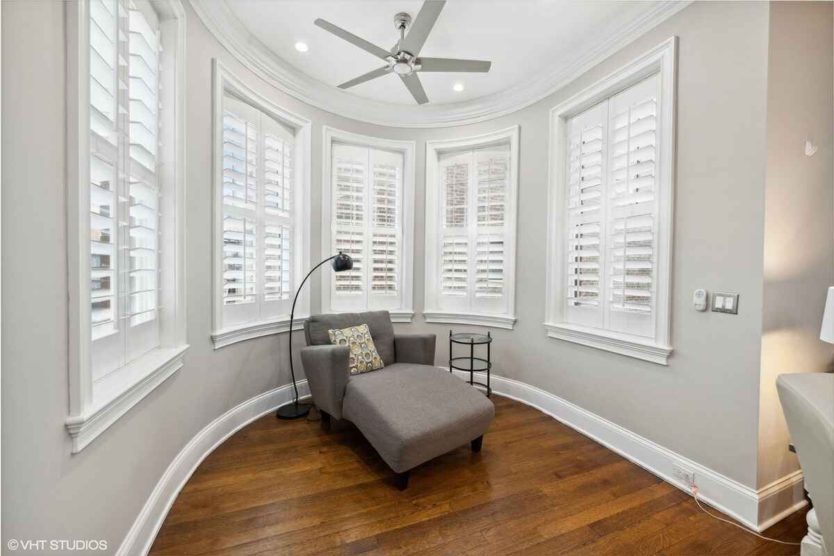 A sitting area with curved walls, multiple windows with shutters, a chaise lounge, a floor lamp, and wooden flooring.