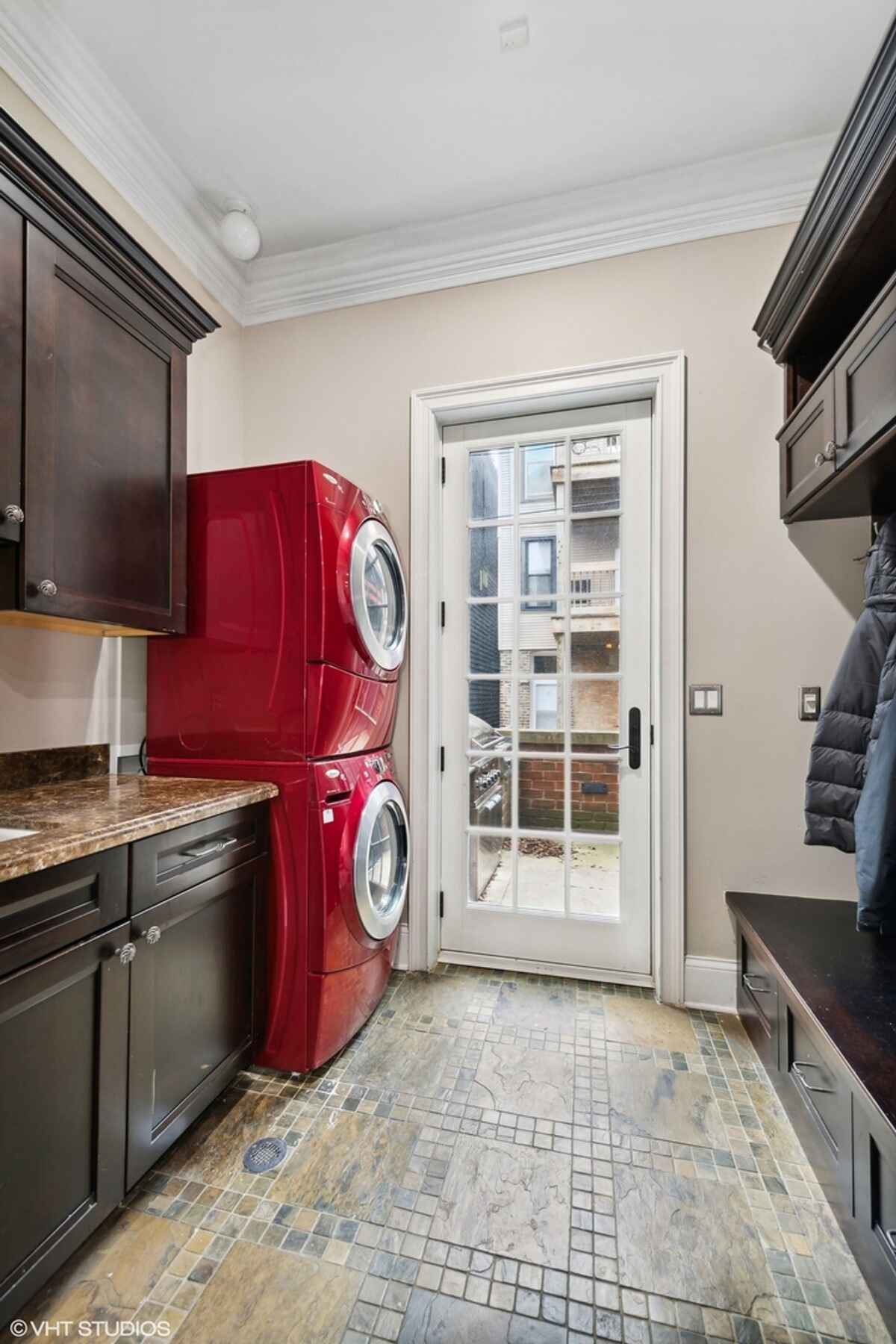 Laundry room contains stacked red washer and dryer units, dark wood cabinets with granite countertops, a tiled floor, and a glass door leading outside.