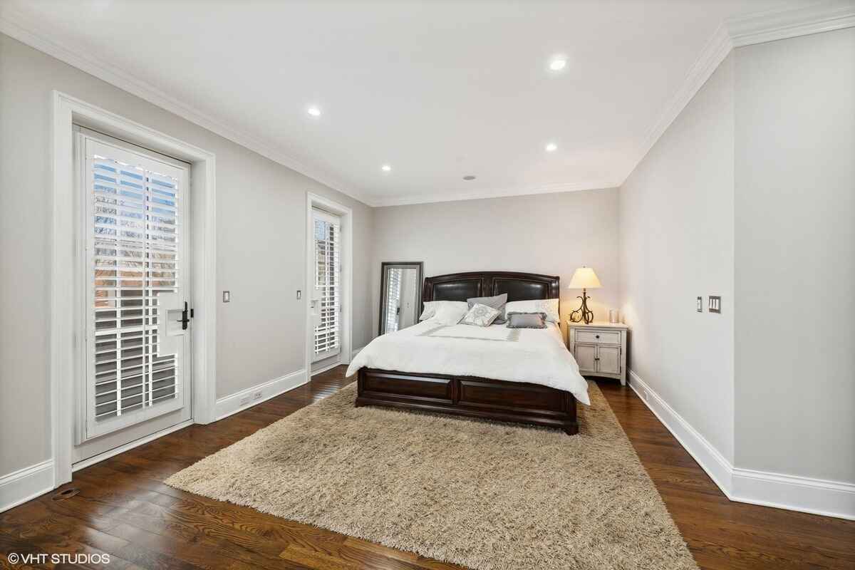 Bedroom features hardwood flooring, a large bed with a dark wooden frame, a soft area rug, and two glass doors with white plantation shutters.