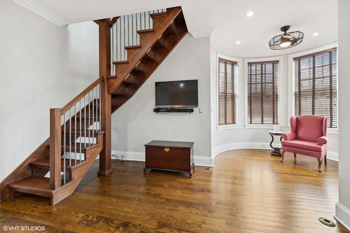 Wooden staircase with metal railings is paired with a hardwood floor and a curved bay window area, featuring a mounted television and a red armchair.