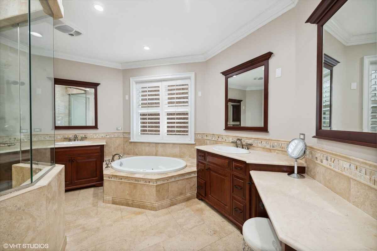 Bathroom includes a built-in bathtub, dual vanities with wood cabinetry, a large mirror, and tiled flooring and walls.