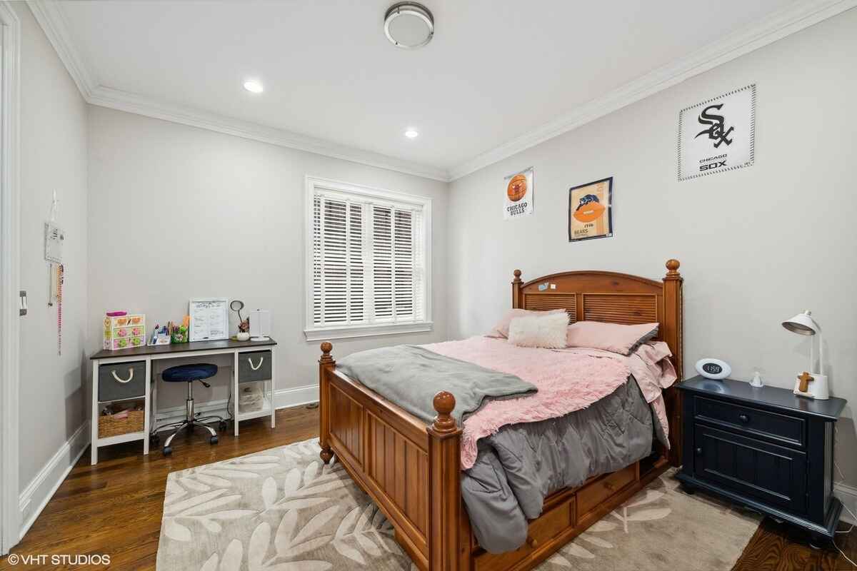 Bedroom features a wooden bed frame with a pink and gray bedding set, a matching nightstand, a small desk with a chair, and wall art above the bed.