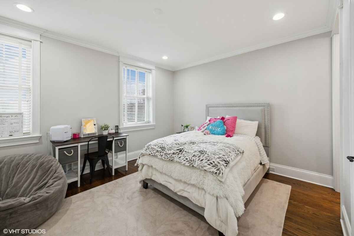 Bedroom features a gray upholstered bed frame with a white bedding set, a small desk with accessories, and two windows with white blinds.