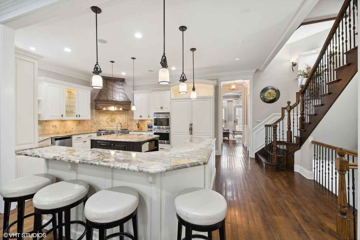 A kitchen with white cabinets, a large island with granite countertops, pendant lighting, stainless steel appliances, and adjacent wooden stairs with wrought iron railings.