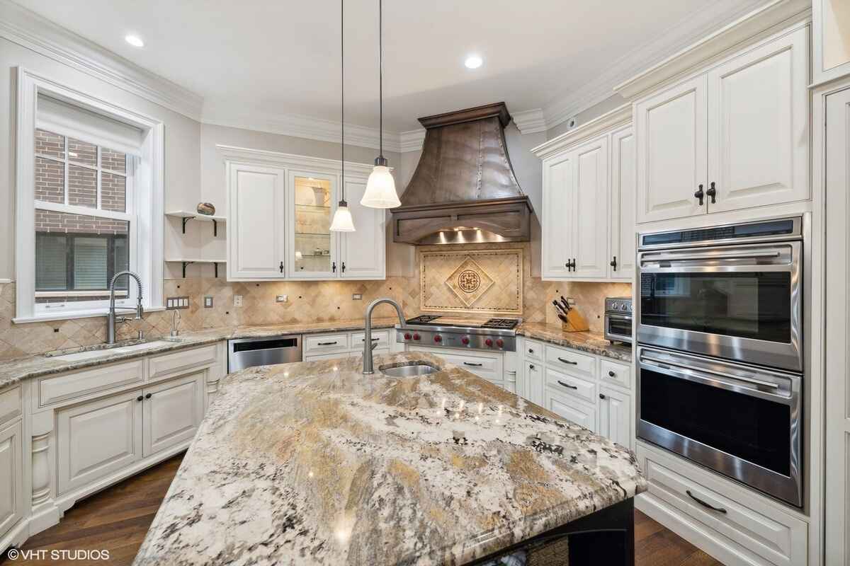 Kitchen features white cabinetry, granite countertops, stainless steel appliances, a large island, pendant lighting, and a decorative backsplash with a central design element.