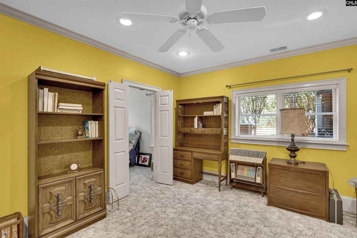 Room has yellow walls, ceiling fan, built-in wooden bookcases and desk, and a window overlooking a yard.