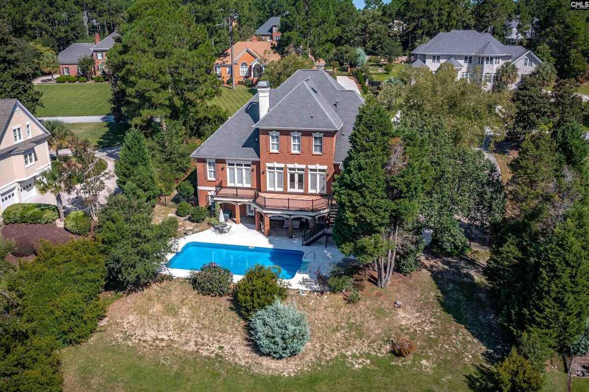 Aerial view shows brick house with a swimming pool, surrounded by trees and other houses.