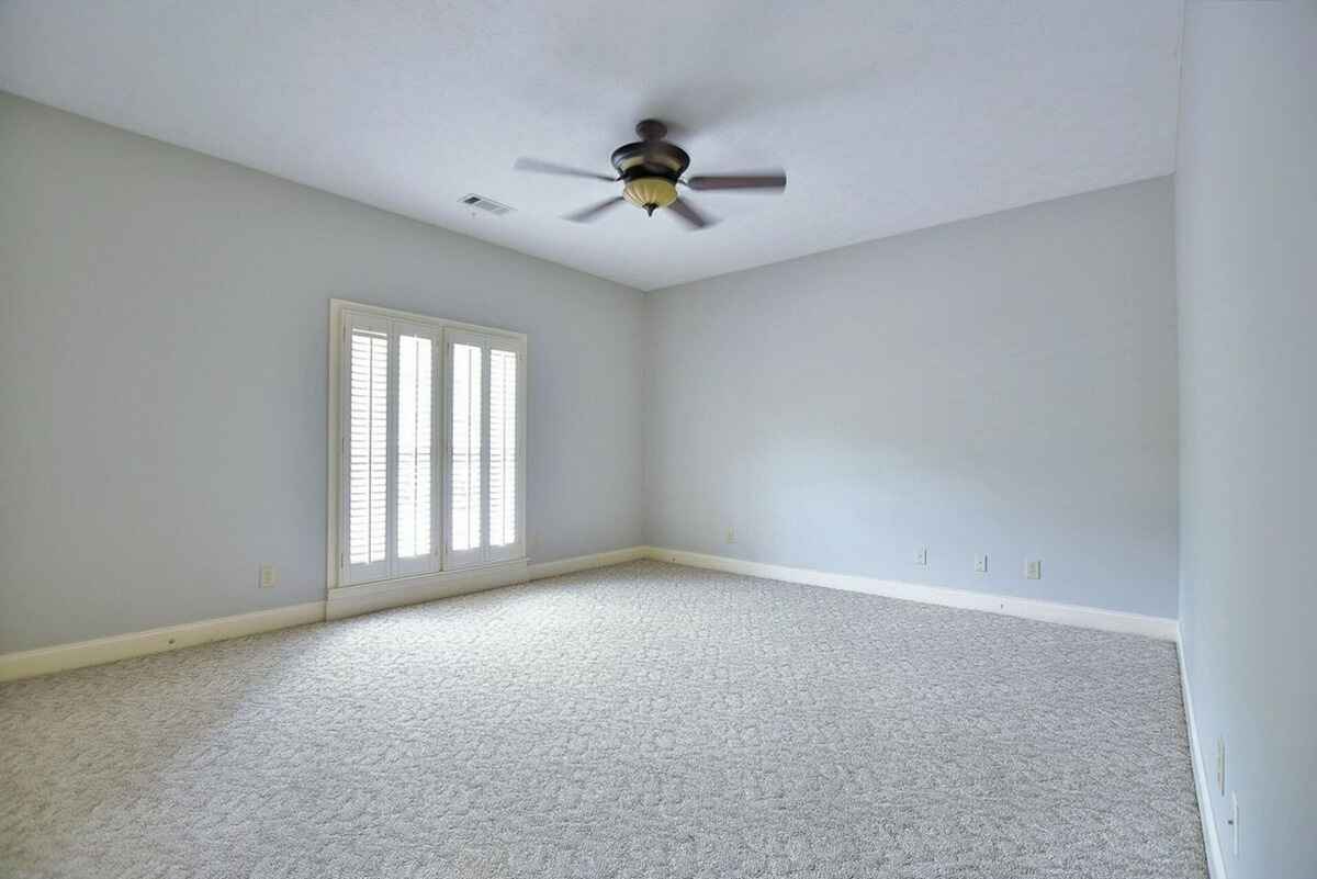 Bright carpeted bedroom showcases a ceiling fan and a large window with shutters.