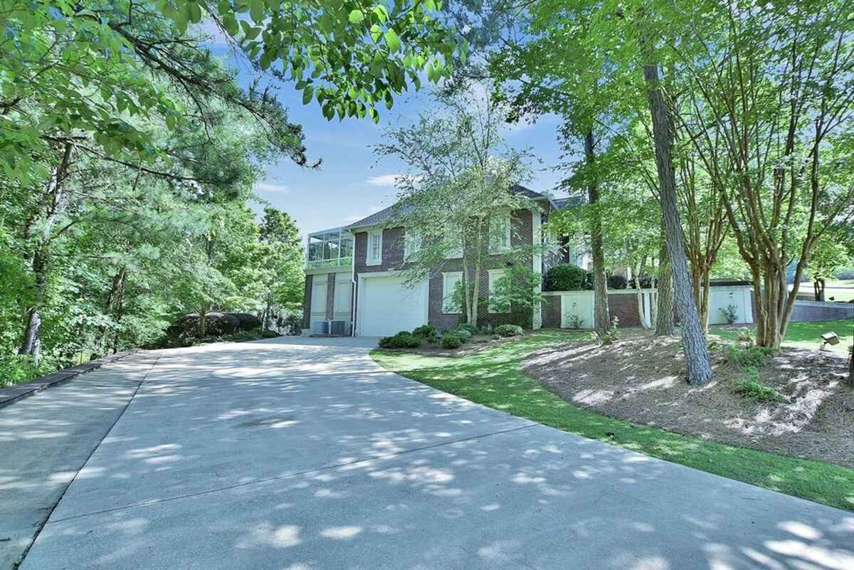 Driveway leads to a two-story brick house surrounded by mature trees and manicured landscaping.