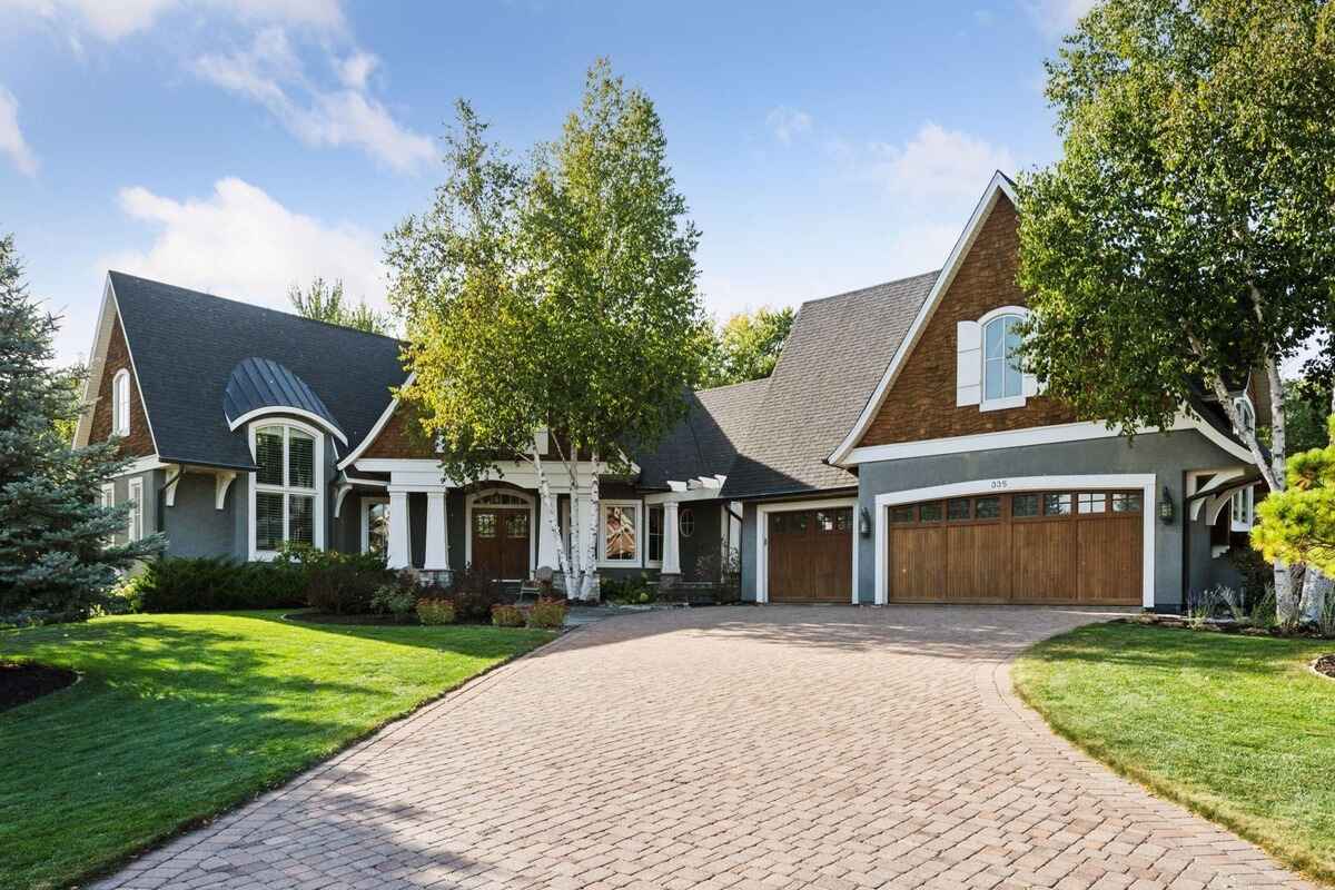 A large home with a paved driveway, wooden garage doors, and trees surrounding the property.