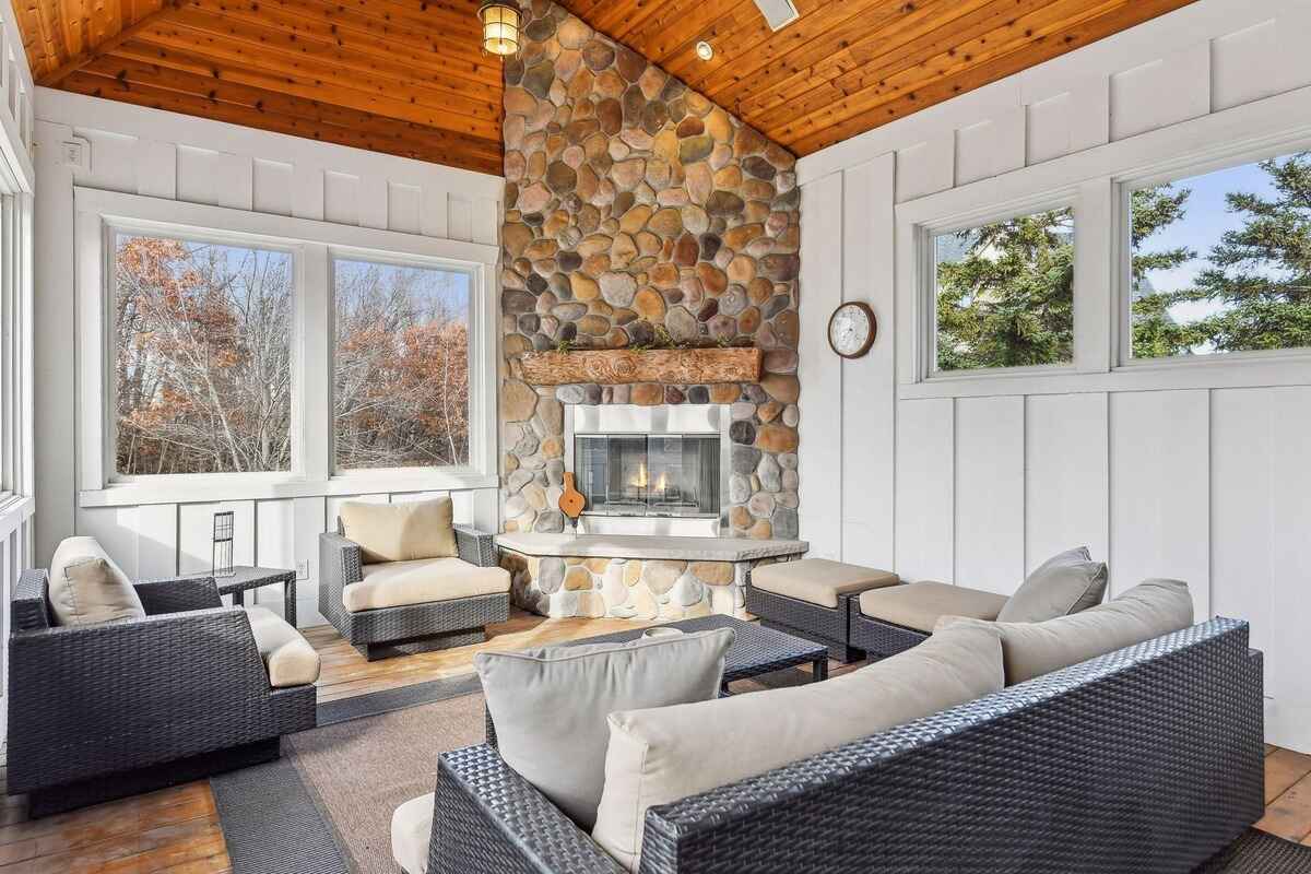 A sunroom features a stone fireplace, wicker furniture, and large windows with a wooden ceiling.