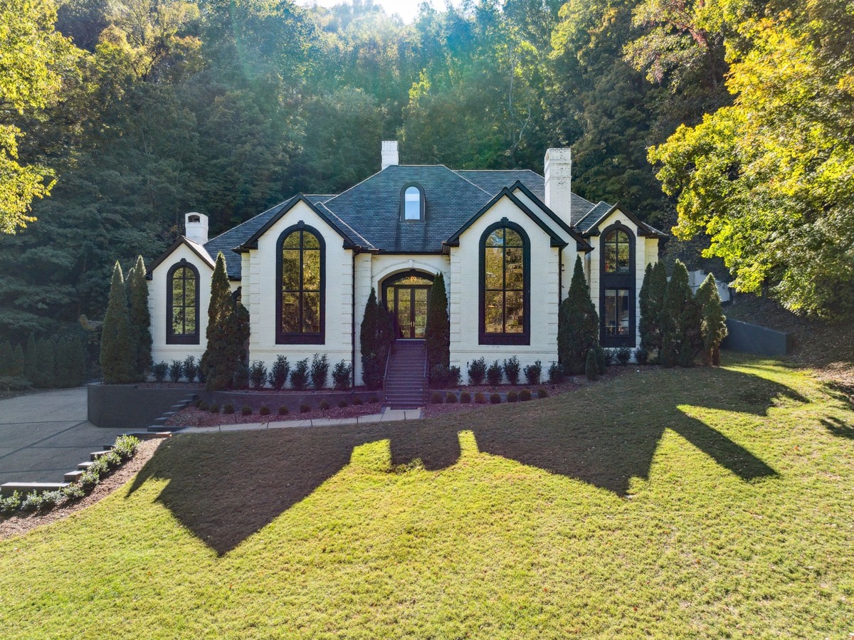 An aerial shot shows a large, white mansion with black trim, situated on a grassy hill surrounded by trees.An aerial shot shows a large, white mansion with black trim, situated on a grassy hill surrounded by trees.