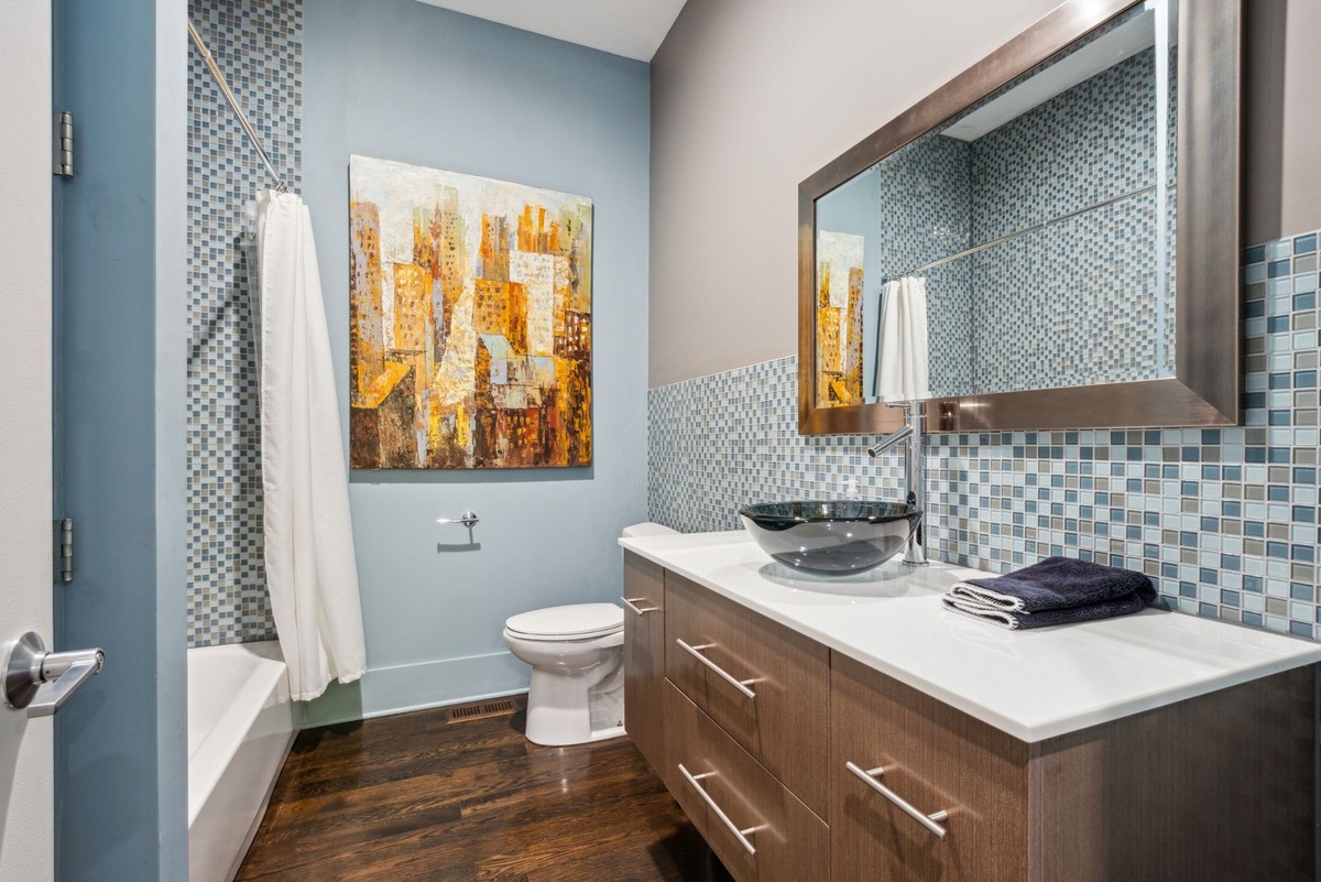 A modern bathroom features a dark wood floor, light blue walls, mosaic tile, a glass sink, and a framed piece of abstract art.