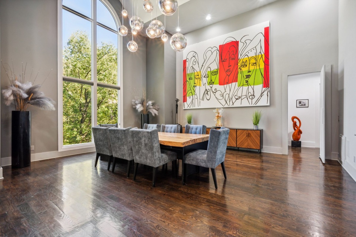 A spacious dining room boasts a long wooden table with gray upholstered chairs, a large abstract painting, and a wall of arched windows overlooking lush greenery.