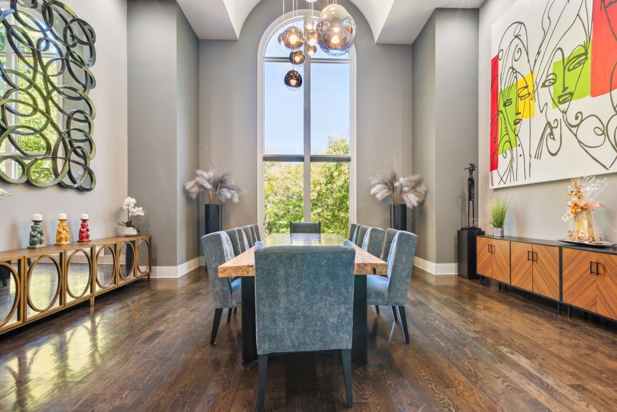 A large, modern dining room features a long wooden table surrounded by teal chairs, a statement art piece, and a large window providing ample natural light.