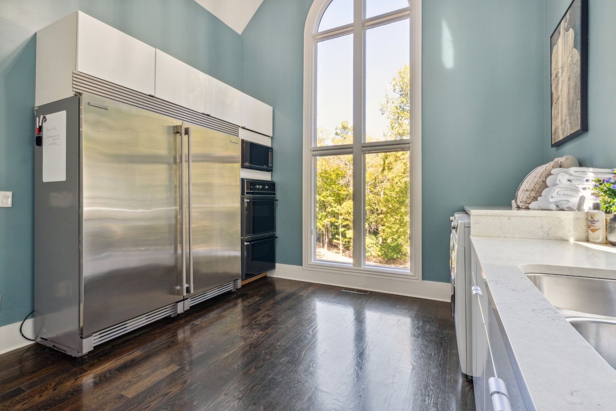 A kitchen features stainless steel appliances, dark hardwood floors, and a large window overlooking a wooded area, with a portion of a laundry room visible in the foreground.