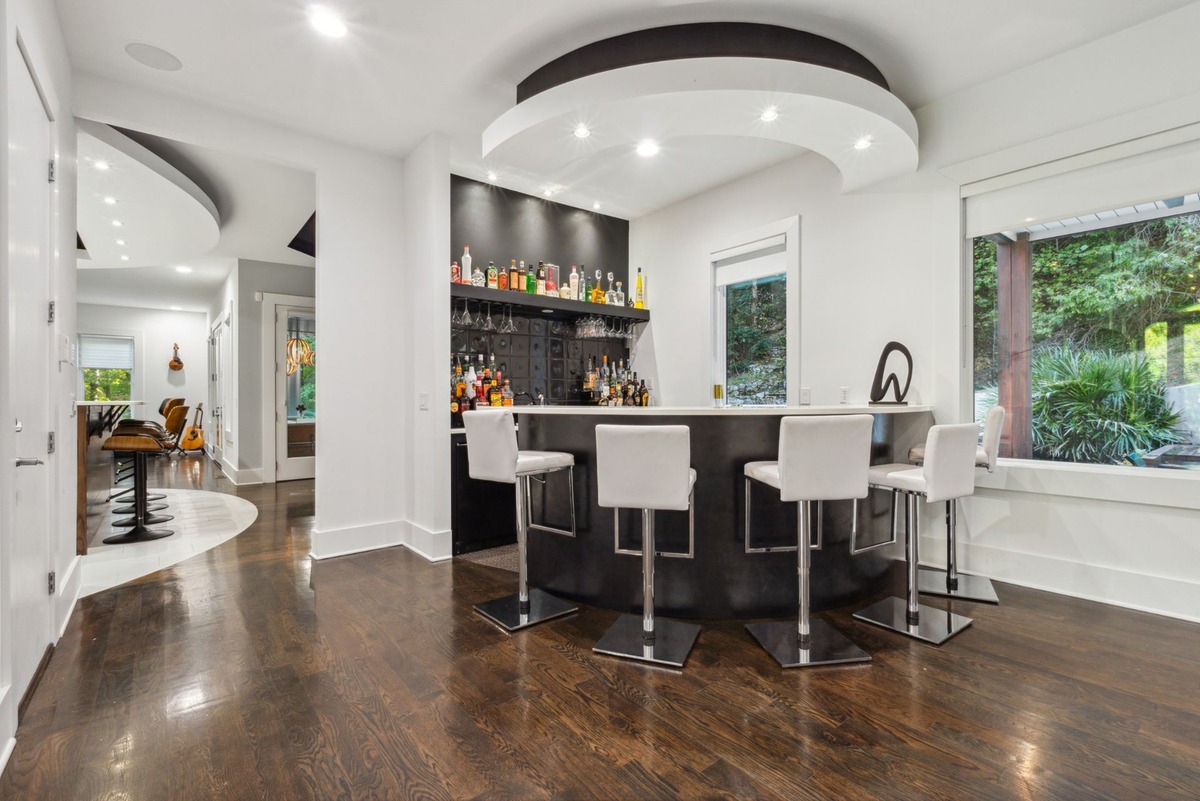 A modern home bar with a curved counter, white stools, and dark wood floors is shown, with a hallway leading to another room visible in the backg