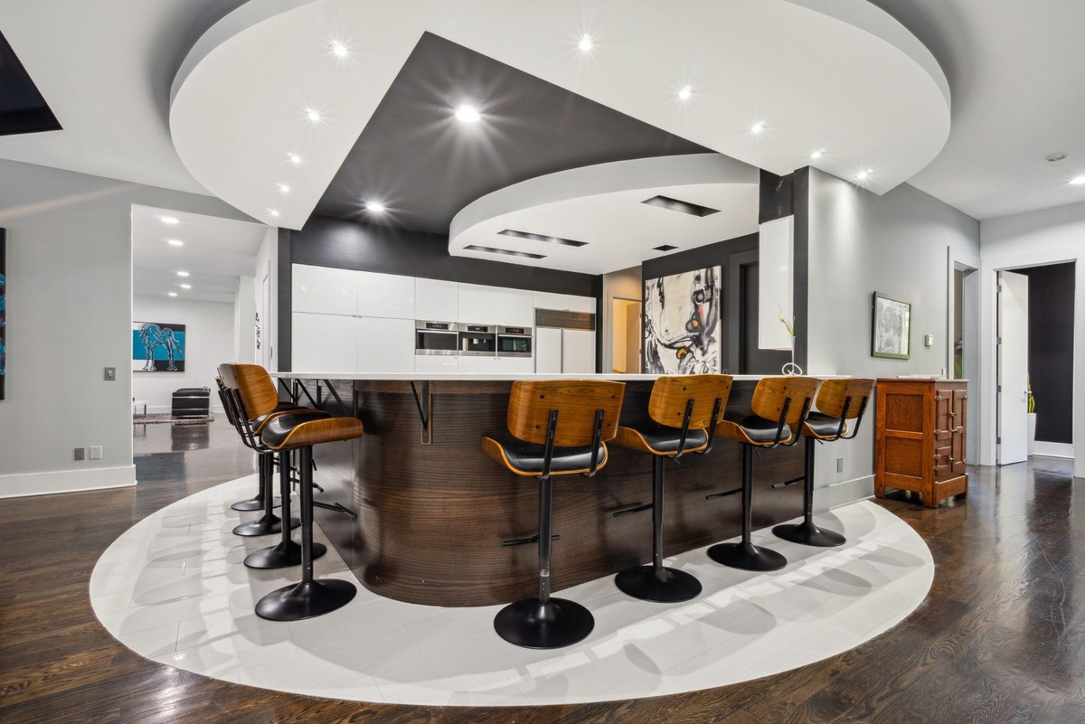 A modern kitchen features a curved island with wooden bar stools and dark hardwood floors, set against a backdrop of white cabinetry and artwork.