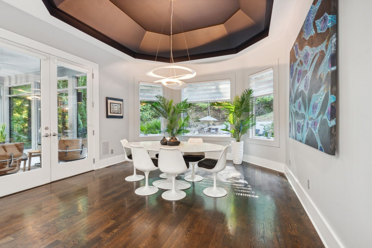 The image shows a modern dining room with a large oval table, white tulip chairs, dark hardwood floors, and large windows overlooking a garden.