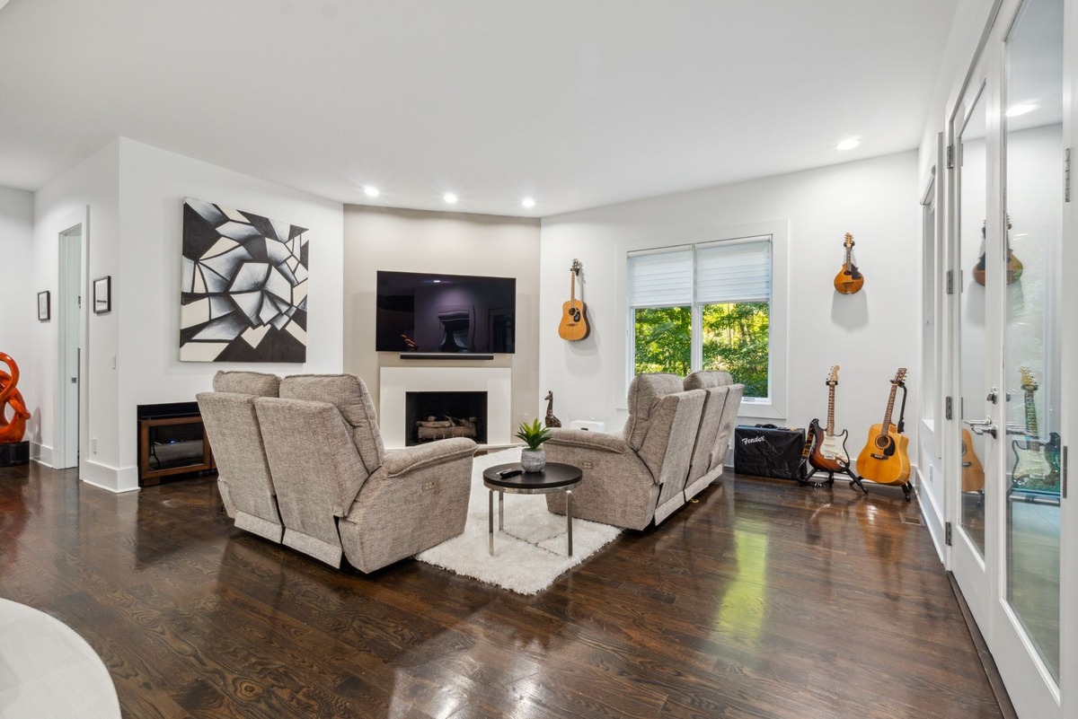 A modern living room features two large reclining chairs facing a fireplace and television, with guitars hanging on the wall and a large abstract painting.