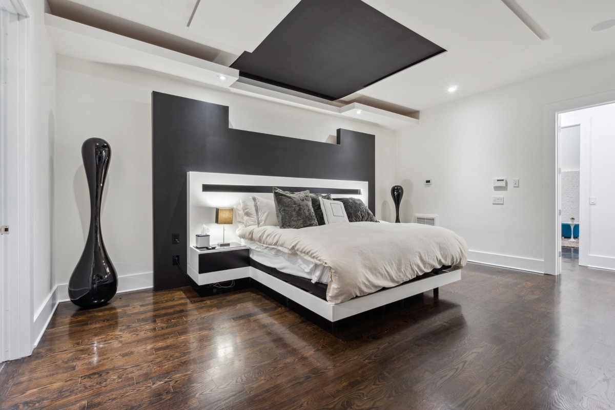 A modern bedroom features a large, white platform bed with a dark headboard against a black accent wall, dark hardwood floors, and a sleek, black floor lamp.